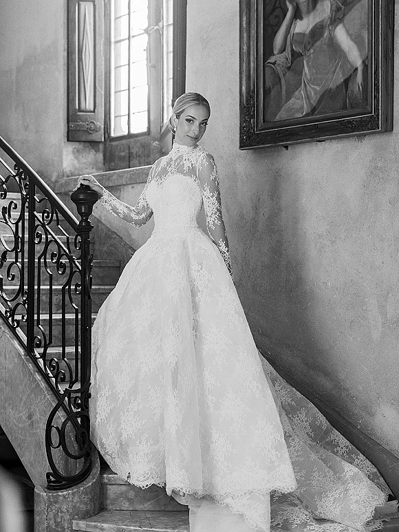 Bridal portrait in a high neck lace ball gown wedding dress with long sleeves, posed on an interior staircase by a tall window