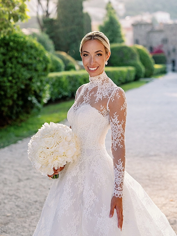 Bridal portrait of a smiling bride in a high neck lace wedding dress holding a white rose bouquet on a gravel path by garden hedges