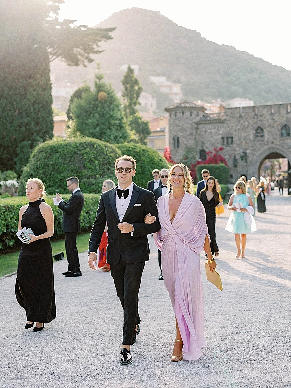 Wedding guest outfit featuring a black tie wedding guest in a tuxedo and bow tie beside a lavender one-shoulder dress in a sunny garden path