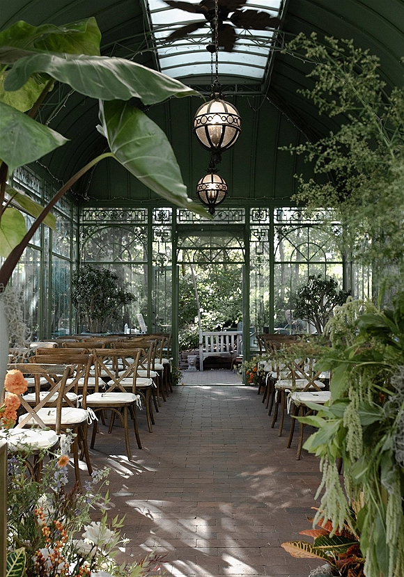 Ceremony setup in a greenhouse wedding ceremony with crossback chairs and white cushions lining a brick aisle with florals and lanterns overhead