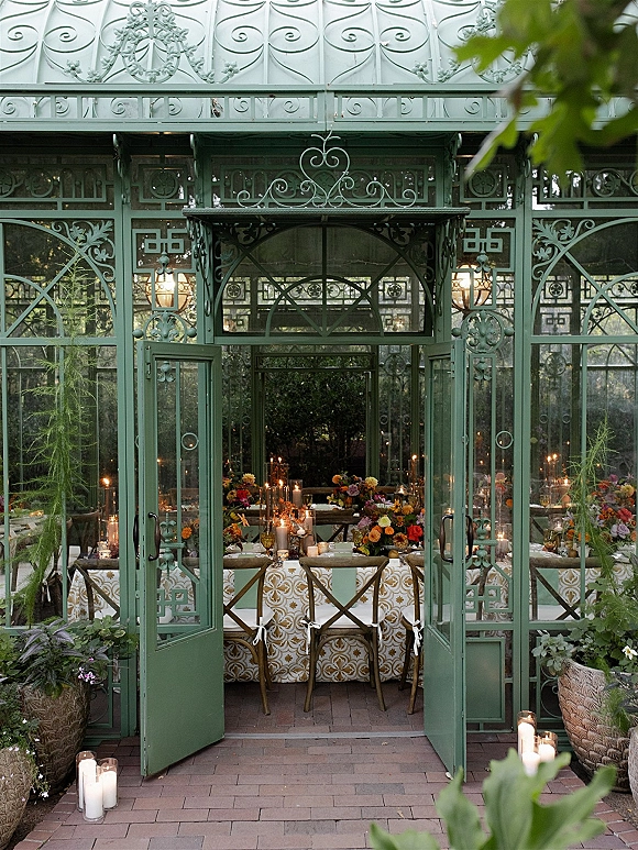 Greenhouse reception decor with long table, patterned tablecloth, floral centerpieces and candles inside green metal glasshouse, doors open to garden greenery