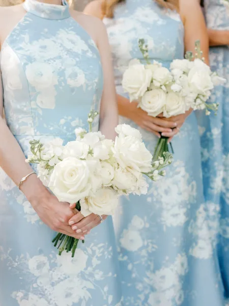 Bridesmaid bouquets of white rose bridesmaid bouquets with greenery held by bridesmaids in blue floral halter dresses, rings visible