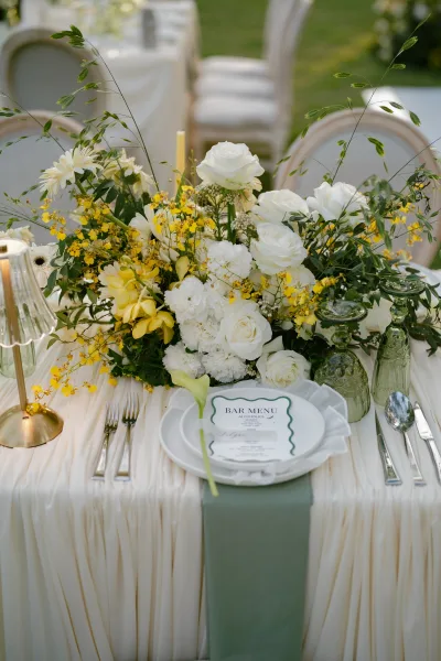 Wedding table setting at a garden reception table with a yellow floral centerpiece, arranged on long tables over a garden lawn.