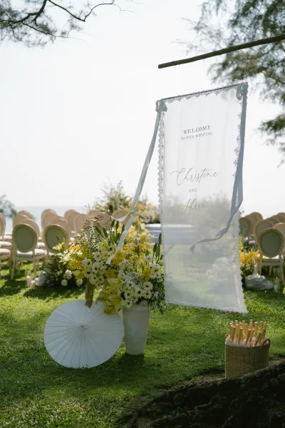 Wedding welcome sign crafted as a fabric welcome sign, framed by yellow and white florals on a grass lawn overlooking the ocean.