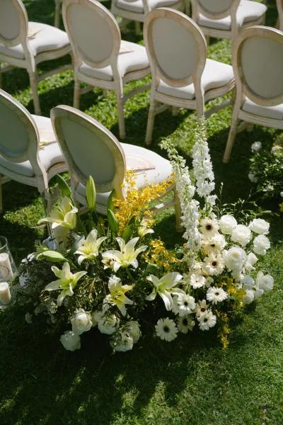 Ceremony aisle flowers line an outdoor ceremony aisle on a grass lawn dappled with sunlight, featuring delicate lilies as focal blooms.
