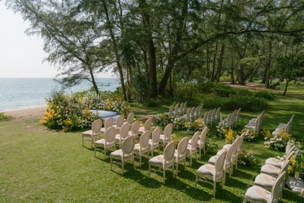 Beach ceremony setup with oceanfront ceremony design, hand fans laid out for guests on a grass lawn facing the ocean and tree-lined shore.