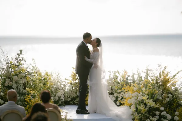 Ceremony kiss captures their first kiss wedding moment, bride in a lace wedding gown and groom, backlit by ocean, sky, and sun.