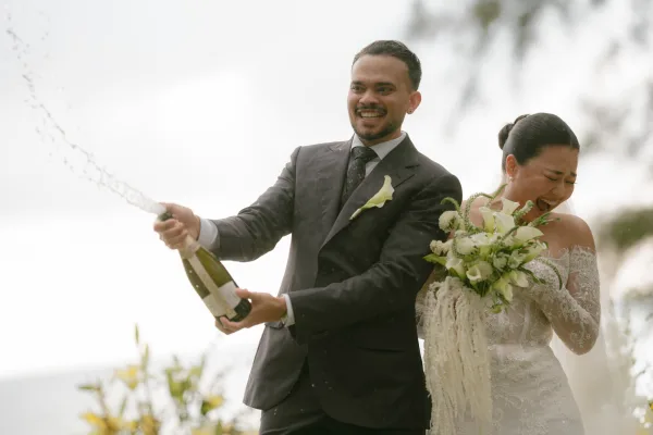 Champagne pop moment with the groom popping champagne as his bride laughs, lace gown shimmering, spray arcs over an ocean backdrop.