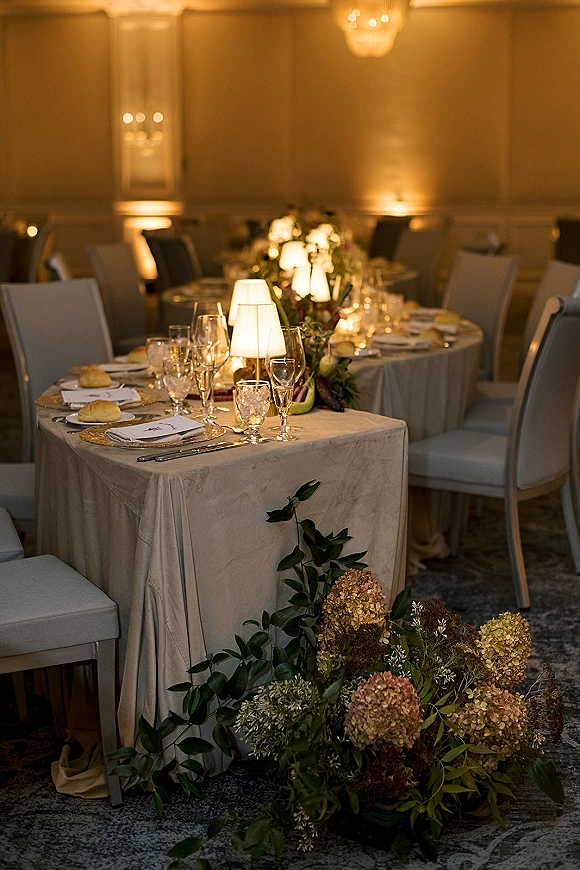 Reception tablescape with a round wedding table setup featuring floral centerpiece, greenery garland, menus, place cards, and gold chargers in a chandelier-lit ballroom