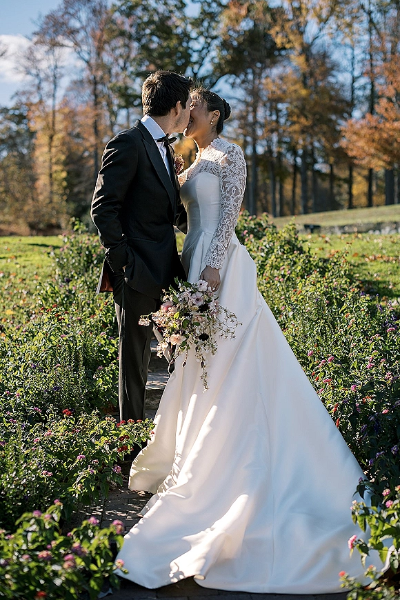 Wedding kiss portrait of bride and groom kissing, bride in lace sleeves holding bouquet beside a garden path under blue sky