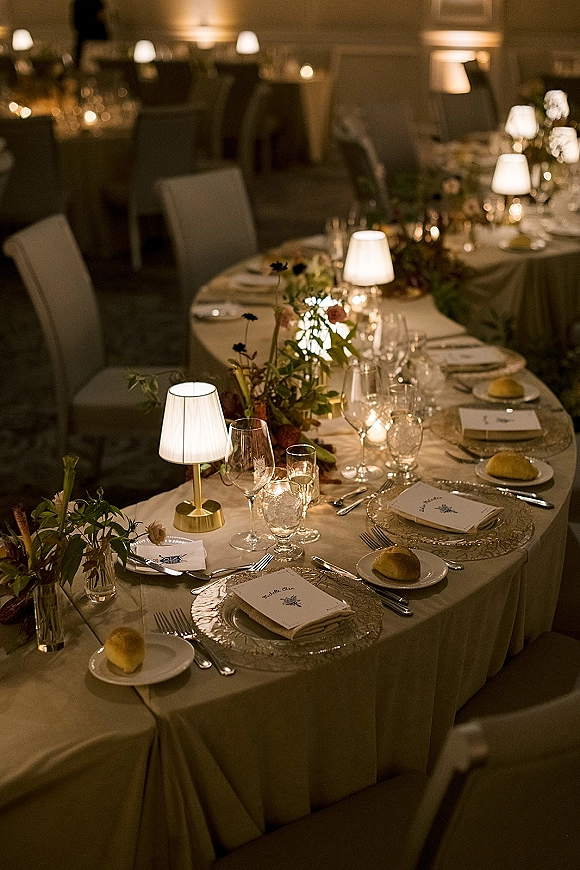 Reception tablescape with wedding head table decor, floral garland and taper candles on white linen, plus table lamps in dim banquet hall