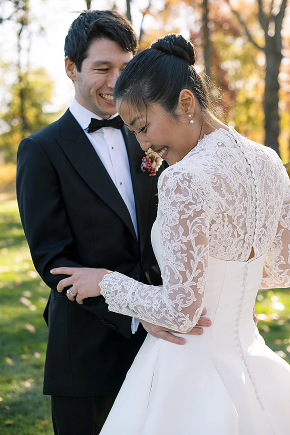 Couple portrait of bride and groom embrace on a sunlit park lawn, her lace long-sleeve dress and pearl earrings beside his tuxedo boutonniere