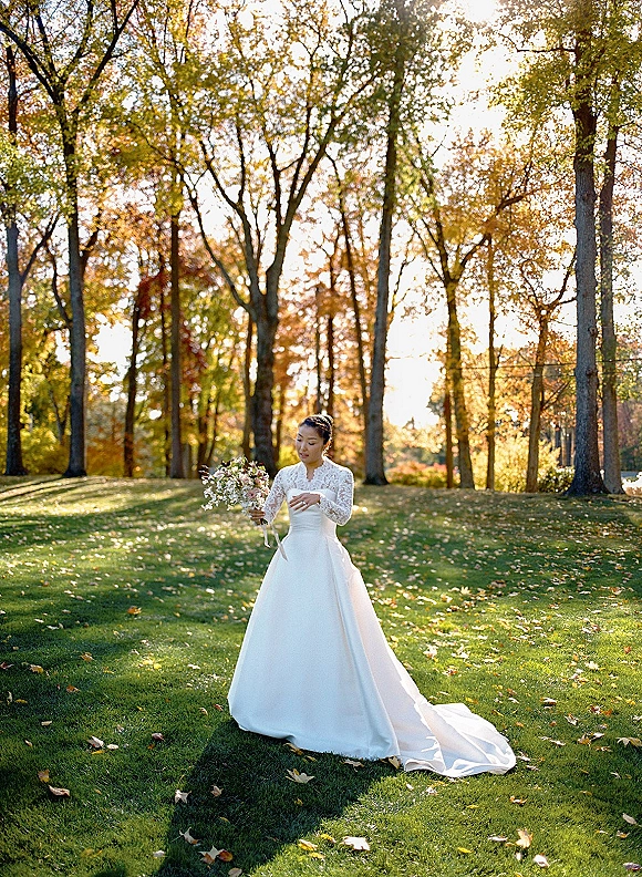 Bridal portrait of a bride looking down holding a bouquet with trailing ribbon, wearing a long sleeve lace dress on a sunlit lawn with autumn trees