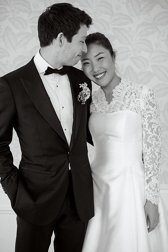 Couple portrait in a black and white wedding portrait style, groom kissing bride’s forehead as she smiles, lace sleeve detail against patterned wallpaper