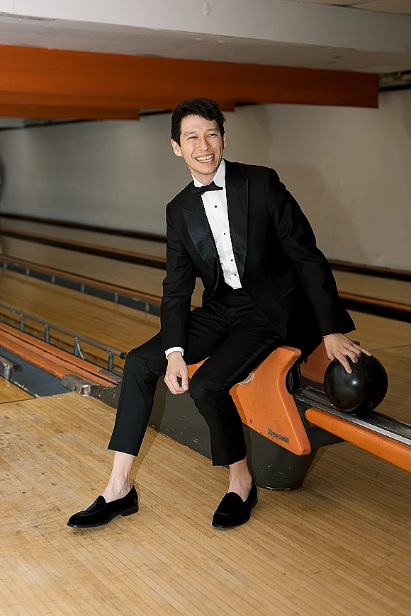 Groom portrait in tuxedo, black tie look as he laughs holding a bowling ball on polished lanes at a bowling alley ball return