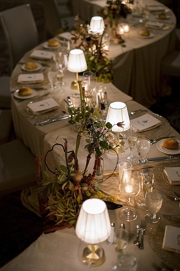 Reception tablescape with wedding head table place settings on white tablecloth, candlelit floral and greenery garland, grapes, and table lamps in dim lighting