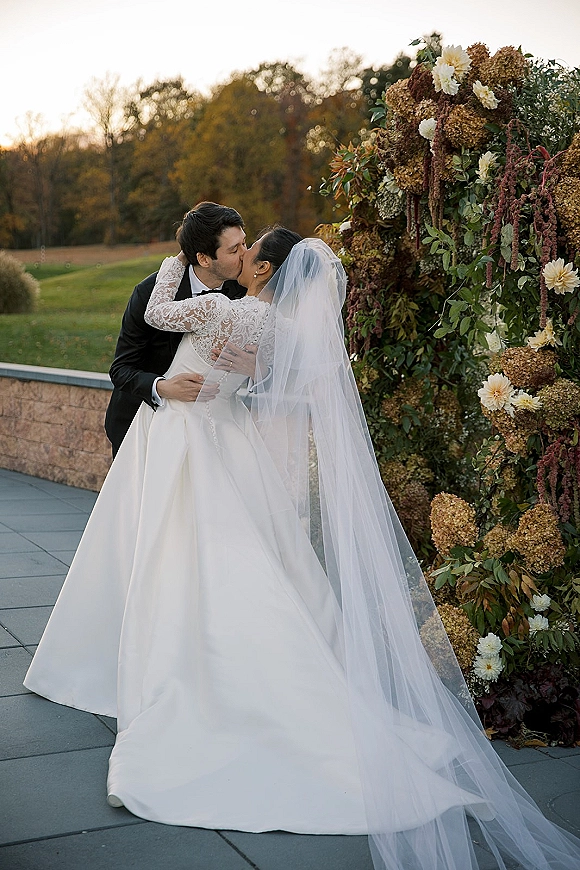 Wedding kiss portrait of bride and groom kissing on an outdoor terrace at sunset, her cathedral veil flowing by a floral wall backdrop