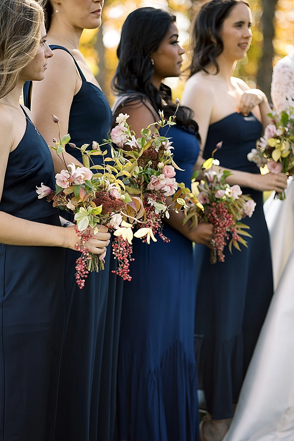 Bridesmaid group in navy bridesmaid dresses holding blush and greenery bouquets, side profile in golden hour light beneath trees