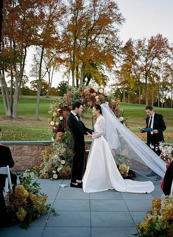 Wedding ceremony moment as bride and groom exchange vows holding hands under a hydrangea floral arch on a stone terrace in autumn