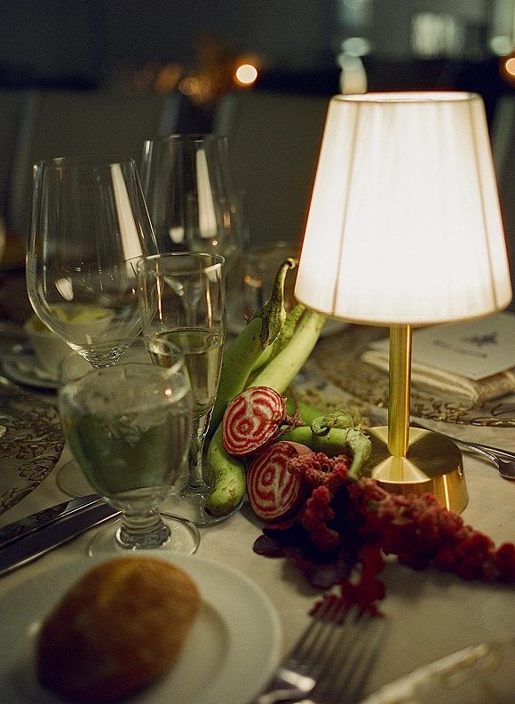 Reception tablescape with a wedding table lamp centerpiece, taper candles, glasses, and patterned linens in a dim room with bokeh lights