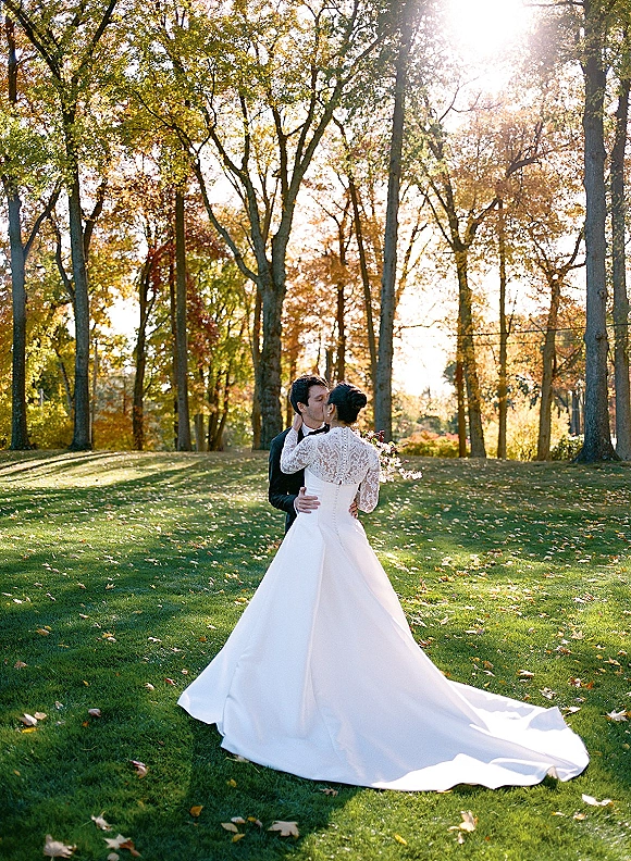 Wedding kiss portrait of bride and groom kissing on a sunlit park lawn, her lace long-sleeve gown with long train and bouquet in hand