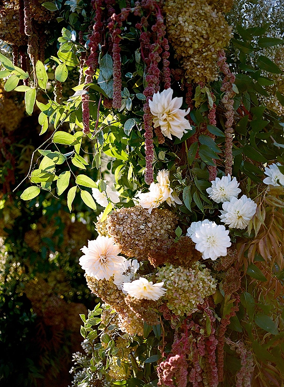 Wedding floral installation with hanging floral installation of white dahlias, hydrangea clusters, and greenery in sunlit garden setting