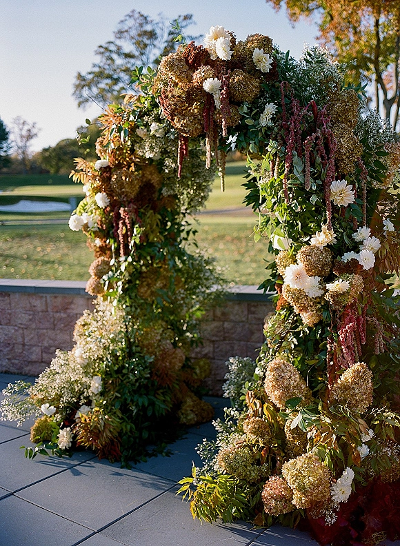 Wedding ceremony arch with a floral ceremony arch of hydrangeas, dahlias, and hanging amaranthus set before a stone wall and lawn