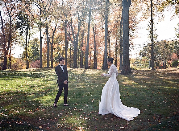 First look moment outdoors as bride in lace long sleeves and long train approaches groom in tuxedo on a sunlit fall park lawn