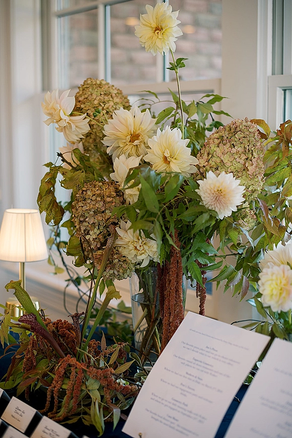 Wedding tablescape with wedding floral centerpiece of dahlias, hydrangeas and greenery in a glass vase, menu and place cards by window panes