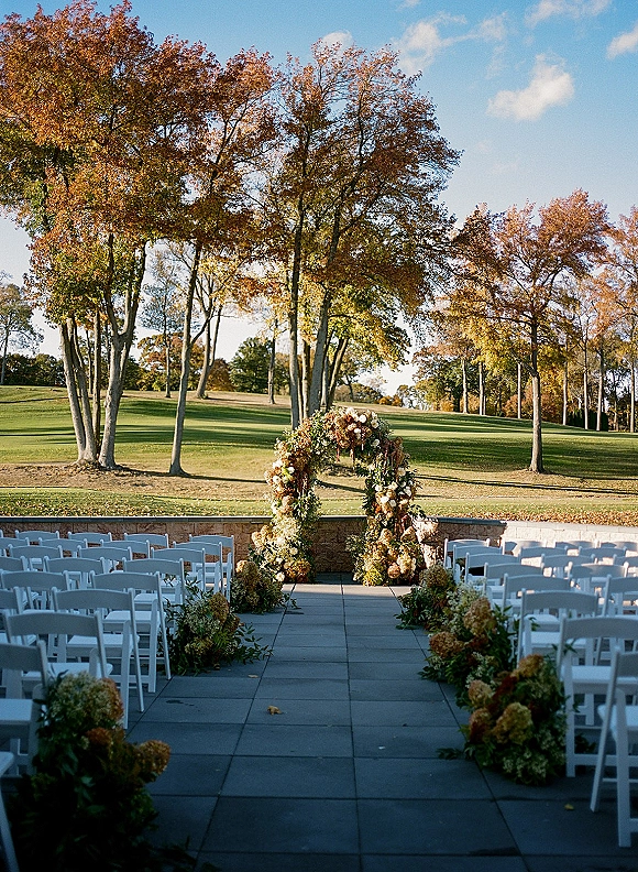 Ceremony setup with a floral arch and aisle florals, white folding chairs on a lawn with trees, blue sky, and stone terrace wall