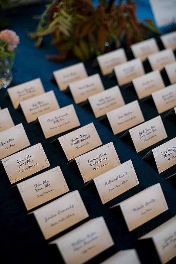 Wedding place cards with calligraphy place cards, ink borders, and greenery on table linen atop a dark tabletop with blurred foliage behind