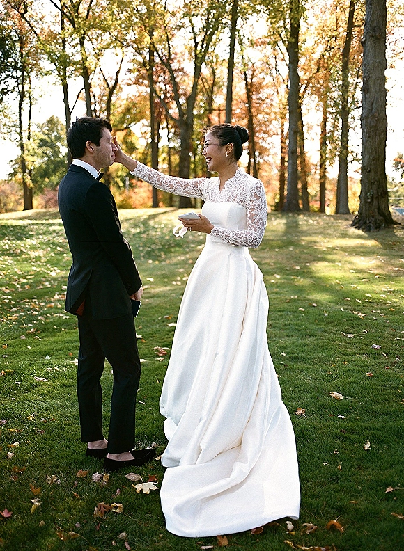 Wedding first look outdoors as bride in lace-sleeved gown touches groom’s face, holding a vow book on a sunlit autumn lawn