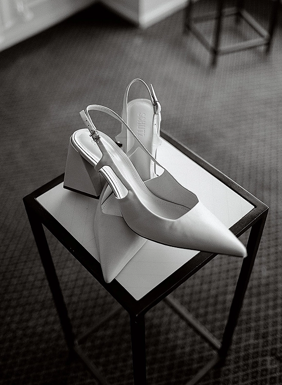 Bridal shoes, white wedding heels with pointed toes and slingback straps resting on a chair above a carpeted floor indoors
