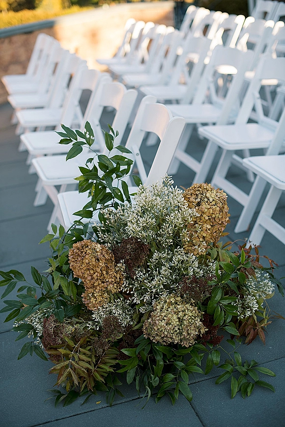 Ceremony aisle decor with wedding aisle flowers, a low hydrangea and greenery arrangement beside white folding chairs on a sunlit patio