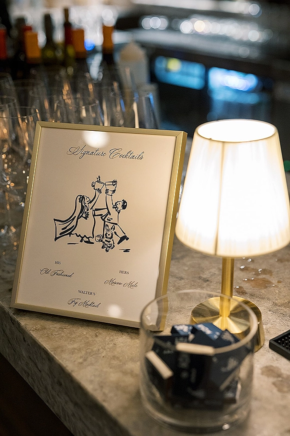 Signature cocktail sign in a gold frame showing an illustrated wedding signature cocktails menu, set on a bar counter with bottles and flutes