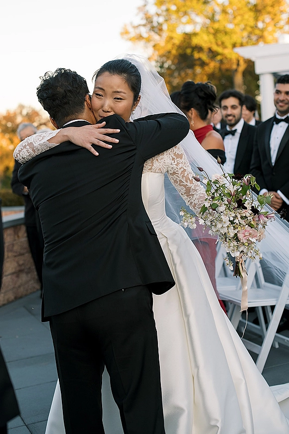 Wedding hug moment as bride hugging groom, bouquet and veil visible, with guests by white chairs near autumn trees and stone wall outdoors