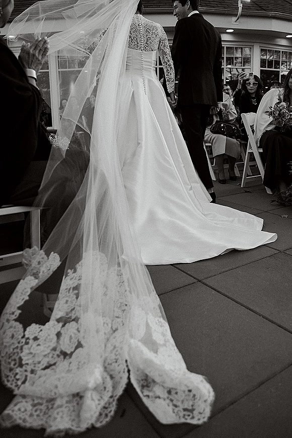 Ceremony recessional as bride and groom walking away hand in hand, veil blowing over lace gown train past clapping guests on patio
