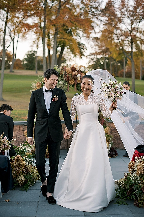 Recessional moment as bride and groom walking hand in hand, her long veil trailing, bouquet in hand on an outdoor terrace aisle