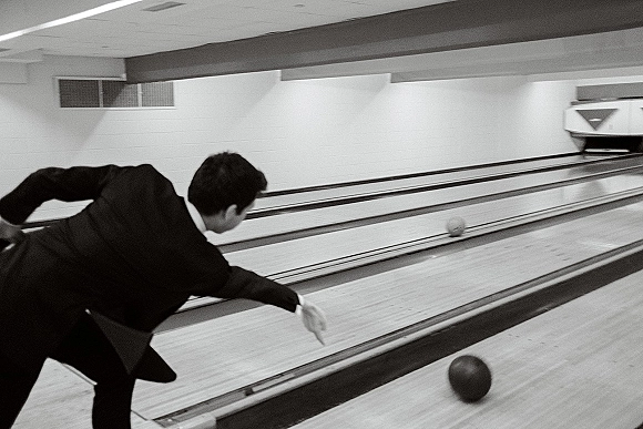 Groom bowling in a tuxedo rolls a bowling ball down an indoor alley lane toward pins under bright ceiling lights