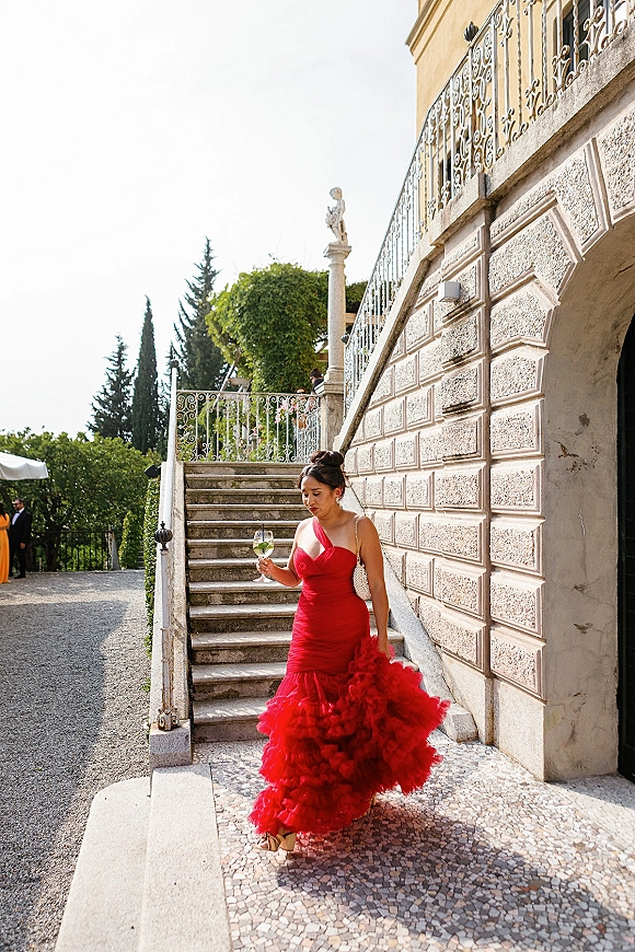 Wedding guest portrait in a red wedding guest dress with dramatic ruffle skirt, holding a wine glass on a stone staircase by ornate iron railing