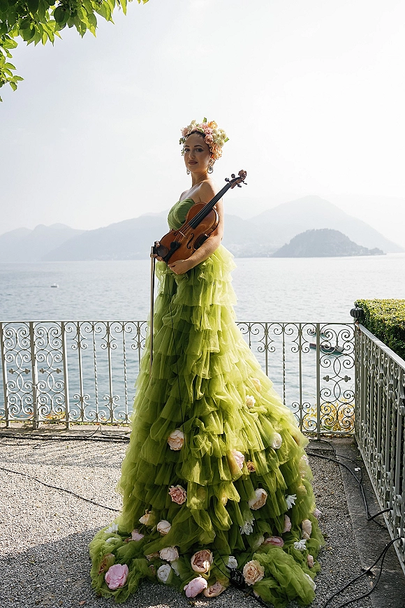 Bridal portrait of a bride in a green wedding dress holding a violin on a terrace, with mountains and a lake behind her