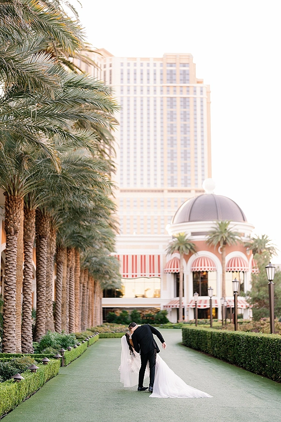 Wedding couple portrait of bride and groom in a dip kiss, her long veil and dress train flowing along a palm-lined garden walkway at a resort hotel