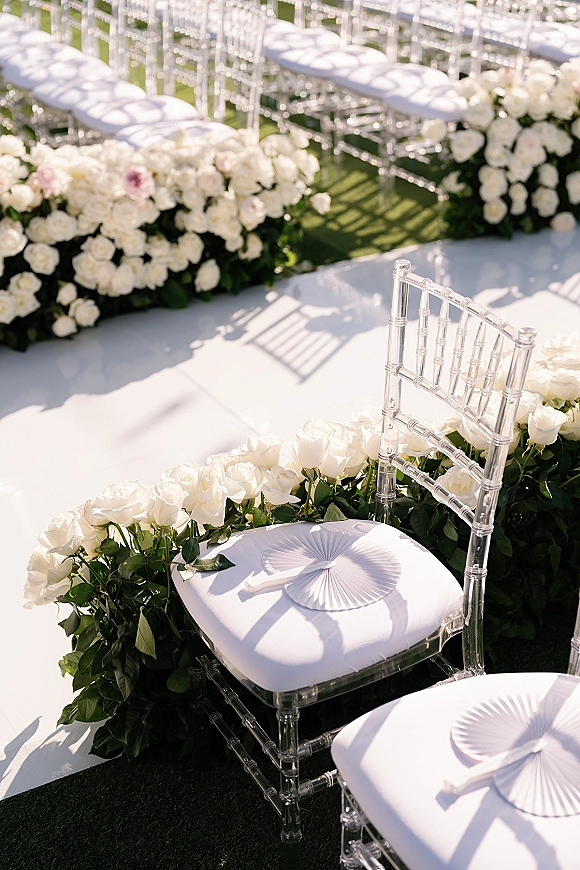 Ceremony aisle decor with white rose aisle flowers lining a white runner, clear Chiavari chairs and paper fans on a sunlit lawn