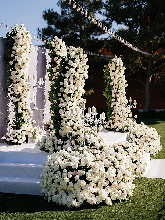 Ceremony floral decor with wedding floral pillars of white and blush roses, greenery and glass candelabras on a white stage outdoors under string lights