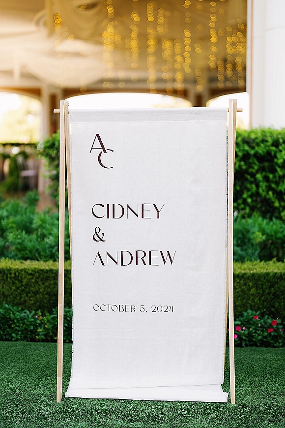 Wedding welcome sign on a wooden stand, fabric linen with monogram and black typography, set by garden hedges with string lights bokeh