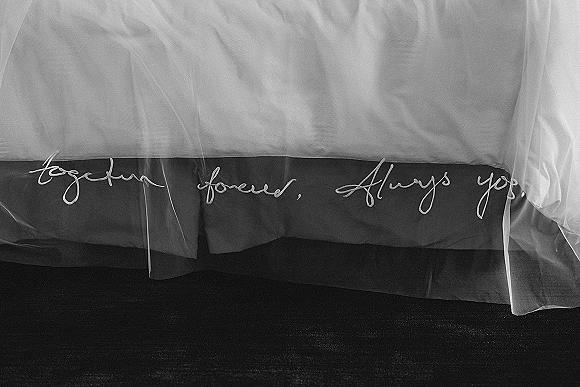 Wedding veil detail with embroidered wedding veil calligraphy stitched into soft tulle, draped over a groom’s suit jacket on carpeted floor