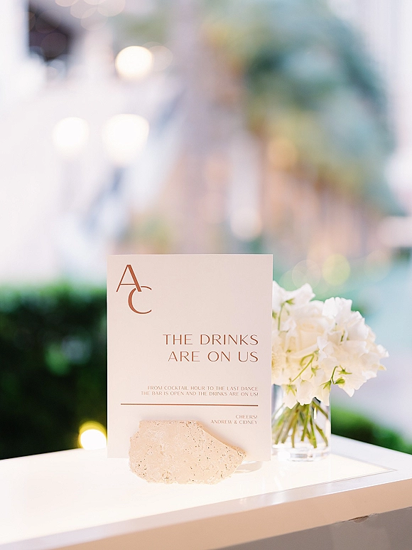 Bar sign displayed in an acrylic holder with monogram initials and white flowers on a bar counter against a hedge wall with string lights