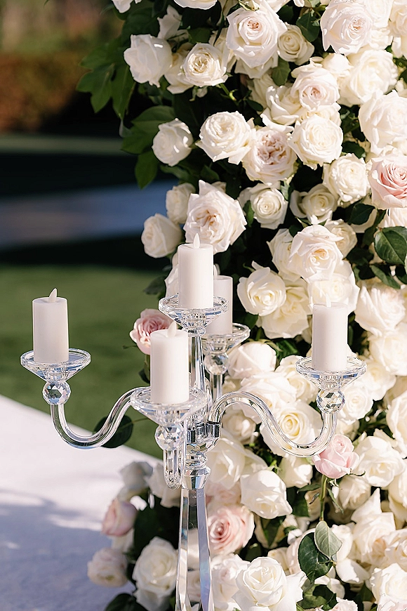 Ceremony floral decor with white rose ceremony flowers, blush roses and greenery around glass candelabra and pillar candles on a lawn