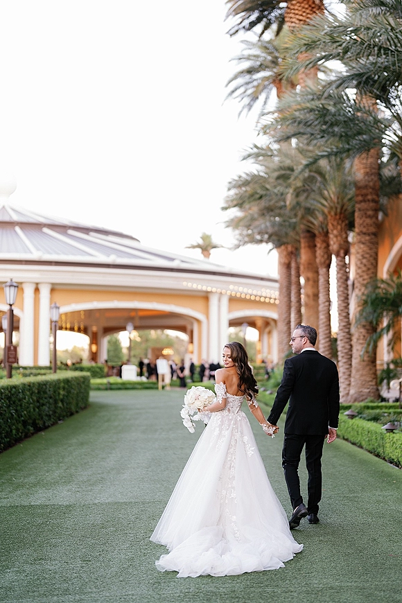 Couple portrait of bride and groom walking away holding hands, bride looking back, lace sleeves and bouquet on a palm-lined resort walkway