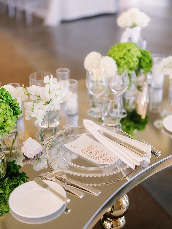 Reception tablescape on a mirrored wedding table with clear charger plates, menu card, white napkin, silver flatware, candles, and hydrangeas in bud vases.